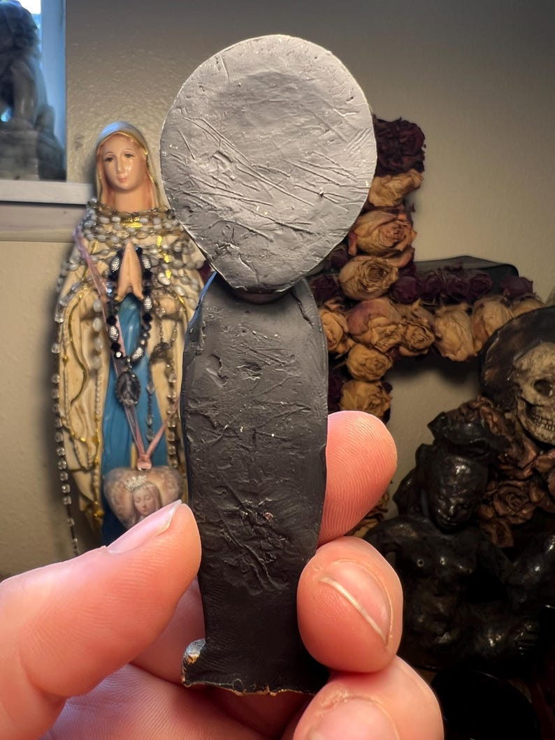 Puede incluir: Una figura hecha a mano, de color gris oscuro, con una cabeza grande y redonda y una superficie texturizada. La figura tiene un cuerpo largo y rectangular y una peque&ntilde;a base. La figura se sostiene en la mano de una persona, con otros objetos religiosos en el fondo.