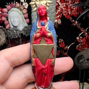 May include: A red and blue figurine of the Virgin Mary with a gold star of David and a gold sun halo. The figurine is holding her hands in prayer and has a blue gemstone in her chest.