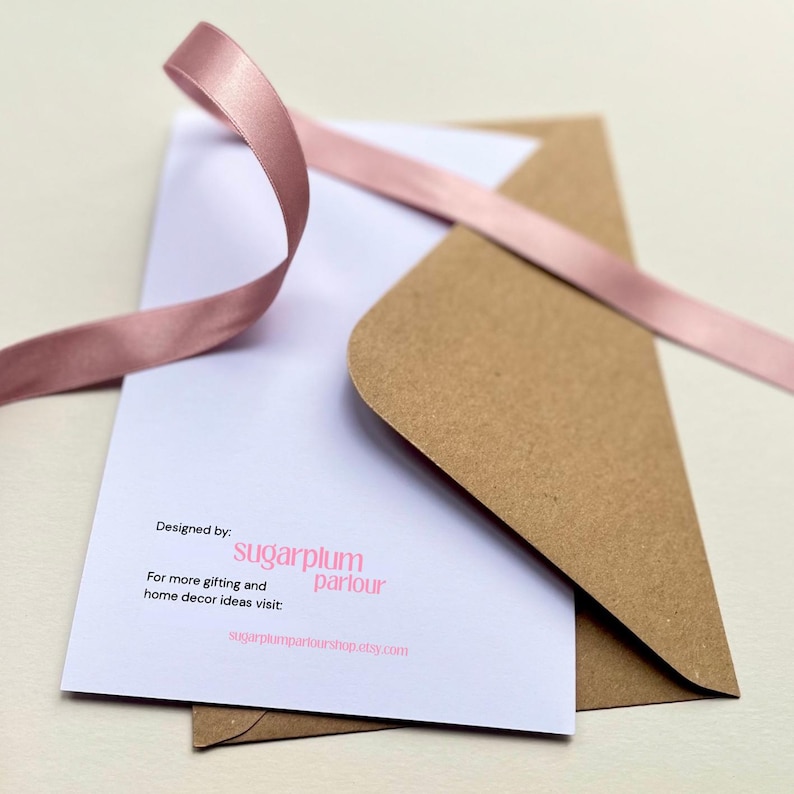 May include: A white card with text and a brown envelope are partially visible, with a pink ribbon draped across them. The card reads "Designed by: sugarplum parlour" and includes a website address.