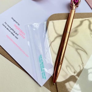 May include: A rose gold pen with a pink gem-shaped top rests on a gold-colored surface. A white card with the text "Designed by: sugarplum parlour" is partially visible. The card also includes the website sugarplumparlourshop.etsy.com.