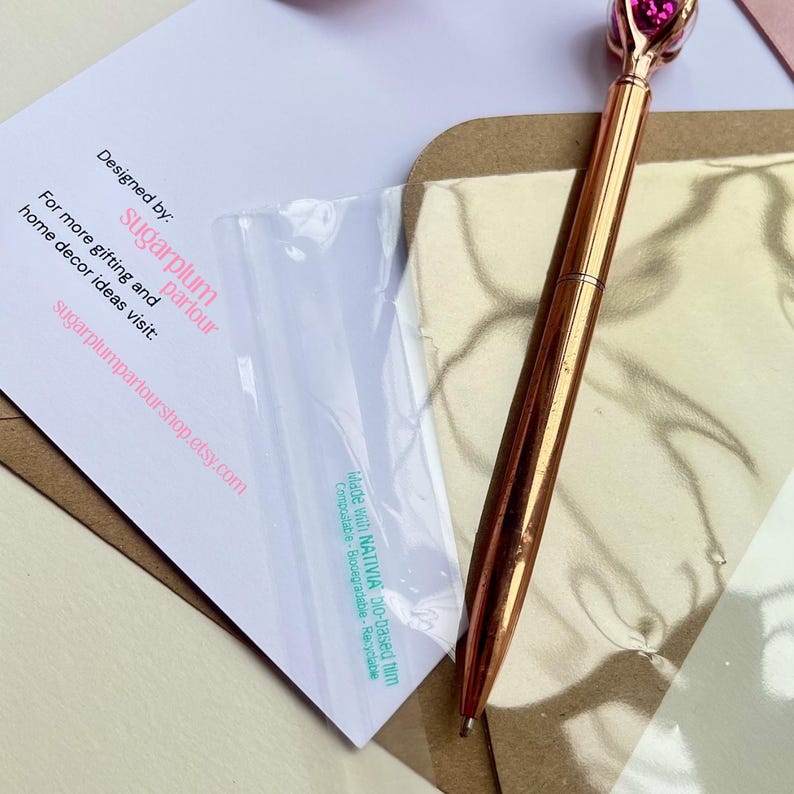 May include: A rose gold pen with a pink gem-shaped top rests on a gold-colored surface. A white card with the text "Designed by: sugarplum parlour" is partially visible. The card also includes the website sugarplumparlourshop.etsy.com.