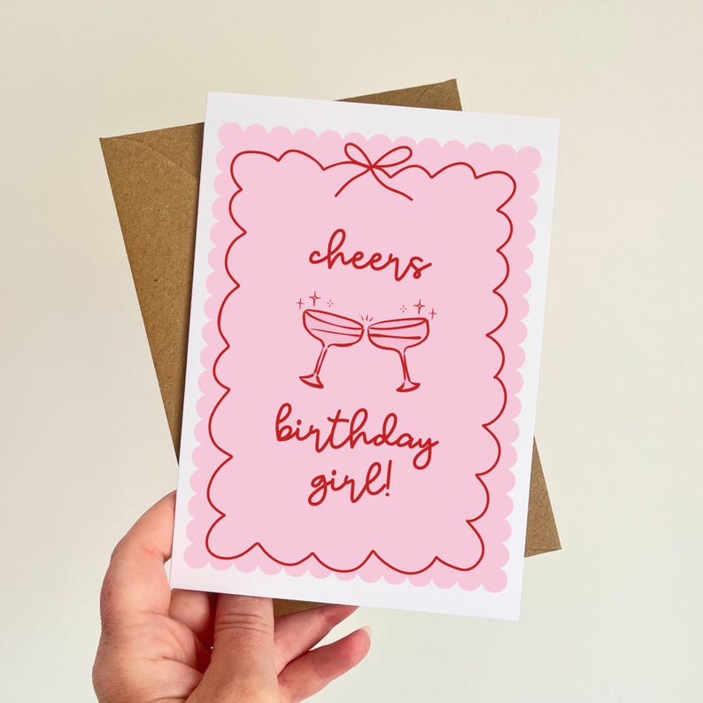 May include: A pink birthday card with a scalloped edge and the words "cheers birthday girl!" in red script. Two champagne glasses are depicted clinking together. The card is held with a brown envelope.