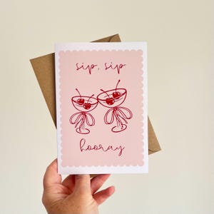 May include: A greeting card with a pink background and a scalloped edge. The card features two red cocktail glasses with cherries, and the words "sip, sip" and "hooray" in red script. A brown envelope is behind the card.
