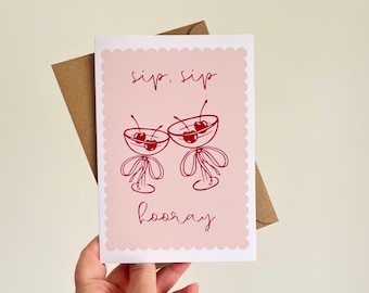 May include: A greeting card with a pink background and a scalloped edge. The card features two red cocktail glasses with cherries, and the words "sip, sip" and "hooray" in red script. A brown envelope is behind the card.