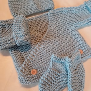 Pode incluir: Um conjunto de bebé de malha azul claro, incluindo um chapéu, casaco e botinhas. O conjunto apresenta um padrão de malha texturizada e é preso com botões de madeira. A cor pastel suave e o design aconchegante sugerem um artigo feito à mão.