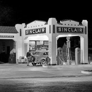 May include: A black and white photo of a Sinclair gas station at night. The station has a white building with the word "Sinclair" in large letters. There is a car being filled with gas at a pump. The sign above the gas pump says "Opaline".