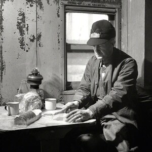 May include: A man wearing a black cap sits at a table with a cup and a loaf of bread. The table is set with a lamp and a glass bottle.