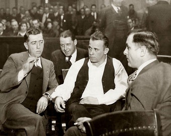 May include: Four men in suits and vests sit in a courtroom. The man on the left is wearing a suit jacket and vest. The man in the middle is wearing a white shirt and a vest. The man on the right is wearing a suit jacket and a vest. The man in the back is wearing a suit jacket and a vest.