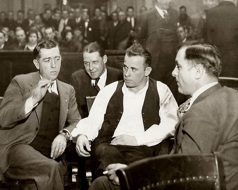 May include: Four men in suits and vests sit in a courtroom. The man on the left is wearing a suit jacket and vest. The man in the middle is wearing a white shirt and a vest. The man on the right is wearing a suit jacket and a vest. The man in the back is wearing a suit jacket and a vest.