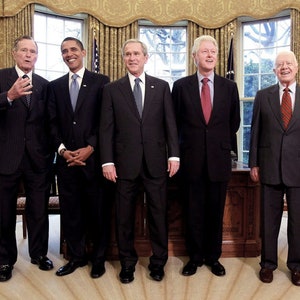 May include: Five former U.S. presidents stand together in a room with white walls and gold trim. They are all wearing dark suits and ties. The presidents are, from left to right, George H.W. Bush, Barack Obama, George W. Bush, Bill Clinton, and George H.W. Bush.