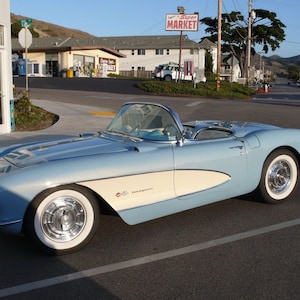 May include: A light blue vintage convertible car with white sidewall tires and chrome hubcaps. The car has a cream-colored side panel and a silver windshield frame. The car is parked on an asphalt road in front of a small town.