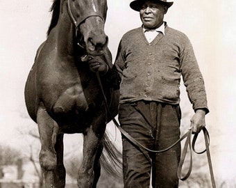 May include: A black and white photo of a man wearing a hat and a sweater, holding a brown horse by the reins.