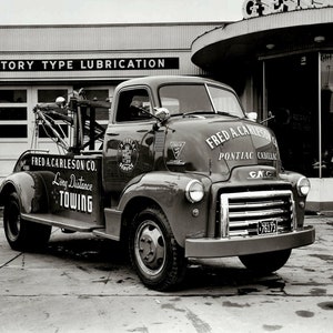 May include: A black and white photo of a vintage tow truck with the words "FRED A. CARLESON CO. Long Distance Towing" on the side. The truck is parked in front of a building with the words "FACTORY TYPE LUBRICATION" above the garage door.