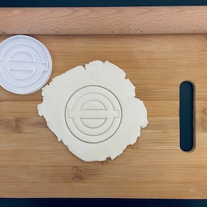 Op de afbeelding: Een witte plastic koekjesvormer in de vorm van het London Underground-symbool op een houten snijplank met een deegroller ernaast. De koekjesvormer ligt op een cirkel van wit deeg.