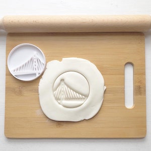 May include: A white plastic cookie cutter shaped like the Golden Gate Bridge on a wooden cutting board with a rolling pin and white fondant.