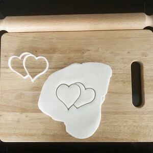 Pode incluir: Uma tábua de cortar de madeira com um rolo e dois cortadores de biscoitos em forma de coração. Fondant branco está na tábua, com duas formas de coração cortadas.