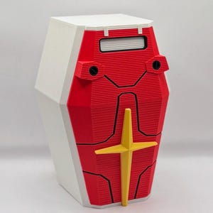 May include: A geometric, red and white container with a yellow cross design. The container has a rectangular shape with a white top and sides, and a red front with black accents. The yellow cross is centered on the red front.