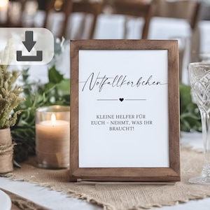 May include: A framed sign with the words "Notfallkorbchen" in elegant script, a heart symbol, and additional text. The sign is in a dark wooden frame, set on a table with a burlap runner, candles, and glassware.