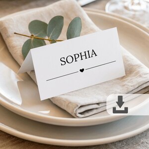 Puede incluir: Una tarjeta de sitio blanca con el nombre "SOPHIA" impreso en negro, una línea horizontal y un pequeño corazón. La tarjeta descansa sobre una servilleta de lino, un plato blanco y está adornada con una ramita de eucalipto.