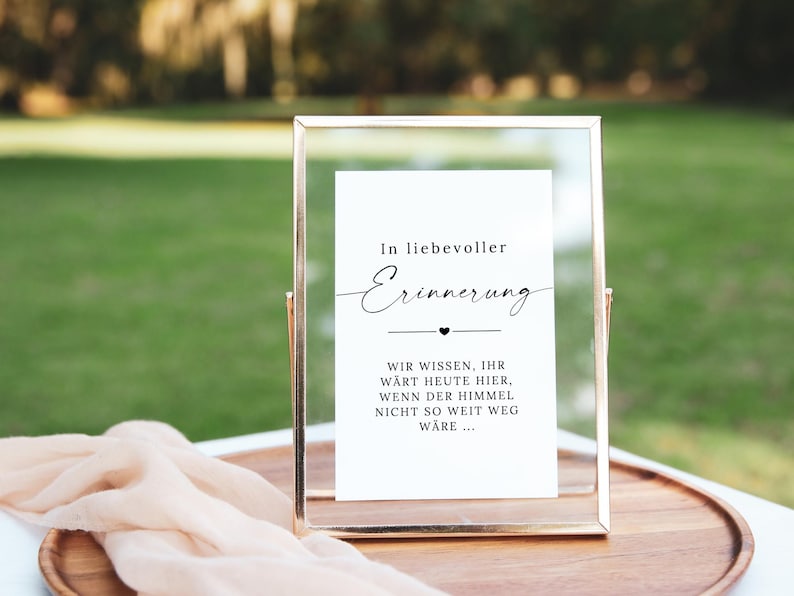May include: A framed sign with gold trim and a white insert. The text on the insert reads "In loving memory" in elegant script, followed by a message. The sign is on a wooden tray with a peach-colored cloth.