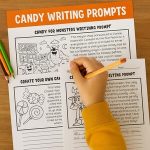 May include: A child's hand writing on a Halloween-themed writing prompt worksheet. The worksheet features illustrations of monsters, candy, and a witch. The title reads "Candy Writing Prompts."
