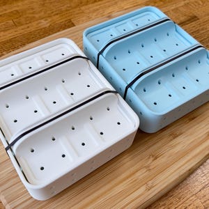 Peut inclure: Deux porte-savons rectangulaires, un blanc et un bleu clair, chacun doté d'une plateforme intérieure surélevée avec de multiples trous de drainage et une bande élastique noire enroulée autour du centre pour maintenir les deux parties.
