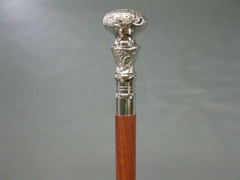 May include: A silver-colored walking cane with a decorative floral design on the handle and a polished wooden shaft. The top of the handle is round and the shaft is a reddish-brown color.