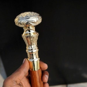 May include: A decorative walking cane with a polished gold-colored metal top and accents. The top features an engraved design. The handle is made of a dark brown wood. The cane is held against a black background.