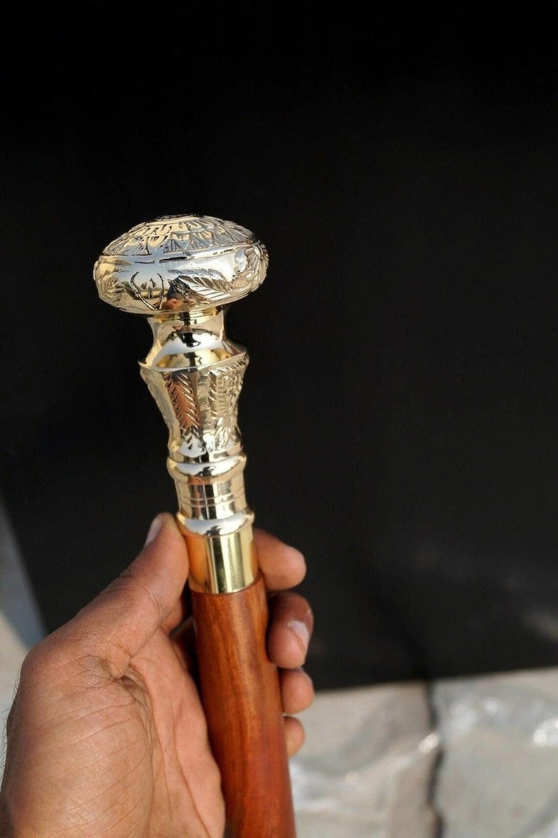 May include: A decorative walking cane with a polished gold-colored metal top and accents. The top features an engraved design. The handle is made of a dark brown wood. The cane is held against a black background.