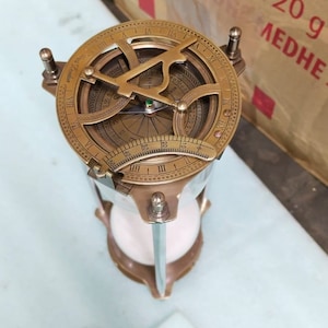 May include: A brass compass with a white hourglass base. The compass face has Roman numerals and markings for degrees. The compass is on a white surface.