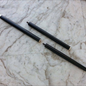 May include: Three-piece black walking cane with gold and silver accents. The cane is disassembled, showing the individual sections. The sections are cylindrical and have a polished finish. The cane is lying on a marble surface.