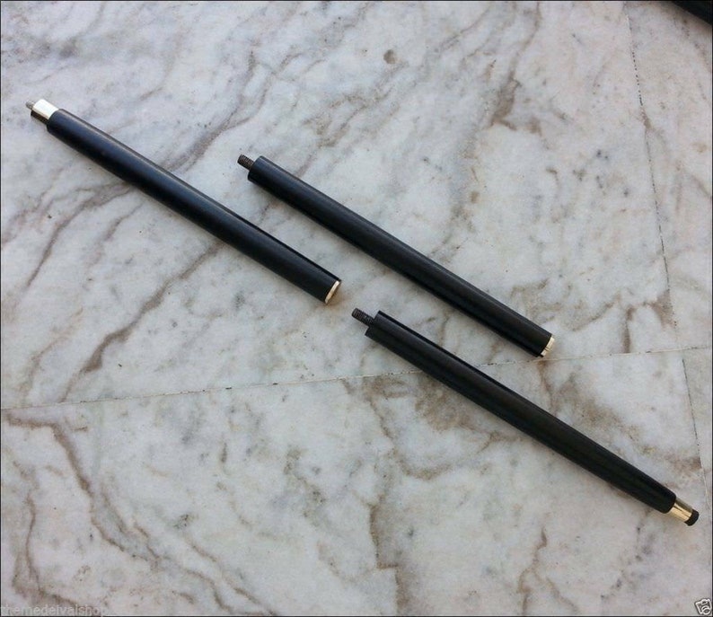 May include: Three-piece black walking cane with gold and silver accents. The cane is disassembled, showing the individual sections. The sections are cylindrical and have a polished finish. The cane is lying on a marble surface.
