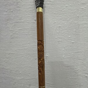 May include: A wooden walking cane with intricate carvings and a bird-shaped handle. The cane features a dark brown finish with gold accents and detailed floral patterns along the shaft. The handle is a dark brown color.