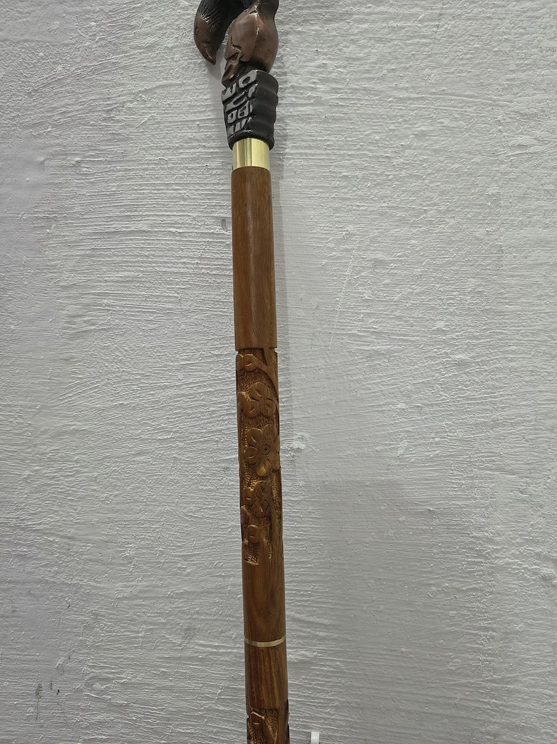 May include: A wooden walking cane with intricate carvings and a bird-shaped handle. The cane features a dark brown finish with gold accents and detailed floral patterns along the shaft. The handle is a dark brown color.