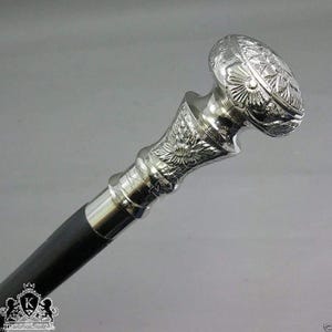 May include: A silver walking stick featuring a detailed floral design on the handle and a black wooden shaft. The handle is oval-shaped with intricate patterns. This decorative cane is a stylish accessory.
