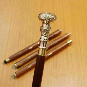 May include: A decorative walking cane with a detailed, ornate brass handle and a dark brown wooden shaft. The handle features intricate floral and geometric patterns. The cane is partially disassembled, with the shaft in multiple sections.