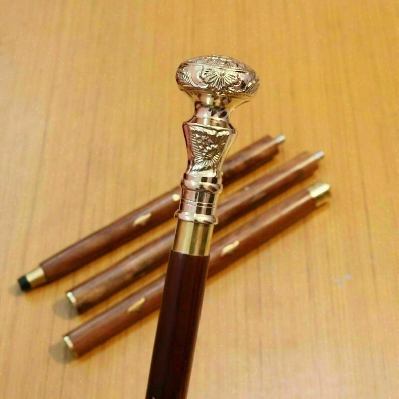 May include: A decorative walking cane with a detailed, ornate brass handle and a dark brown wooden shaft. The handle features intricate floral and geometric patterns. The cane is partially disassembled, with the shaft in multiple sections.