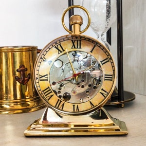 Puede incluir: Un reloj dorado de estilo vintage con números romanos y engranajes internos visibles. El reloj tiene una esfera redonda y se apoya sobre una base rectangular. Un asa dorada está en la parte superior. A la izquierda hay una jarra de latón con un diseño de ancla.