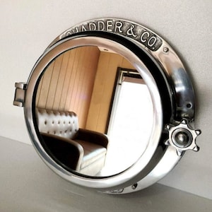May include: A round, silver-colored mirror with a nautical design. The mirror has a thick, metallic frame with the text "QUADDER & CO." embossed on the top. It features a hinged latch and a star-shaped knob. The mirror reflects a room with a brown leather couch.