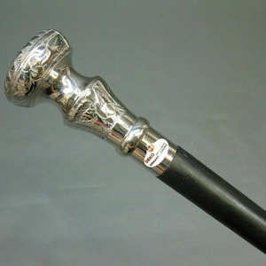 May include: A silver-colored walking cane with a decorative floral design on the handle. The handle is silver, and the shaft is black. The cane has a label that reads "DEKO GERMANY ZWICKAU".