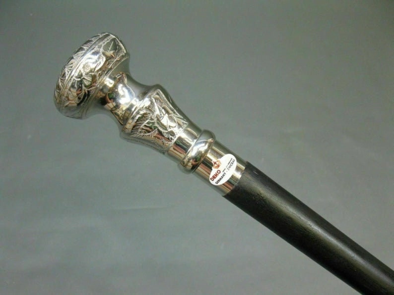 May include: A silver-colored walking cane with a decorative floral design on the handle. The handle is silver, and the shaft is black. The cane has a label that reads "DEKO GERMANY ZWICKAU".