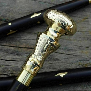 May include: A close-up of a black walking cane with a gold-colored handle. The handle has an ornate design with floral patterns. The cane's shaft is black with gold accents. The cane is resting on a wooden surface.
