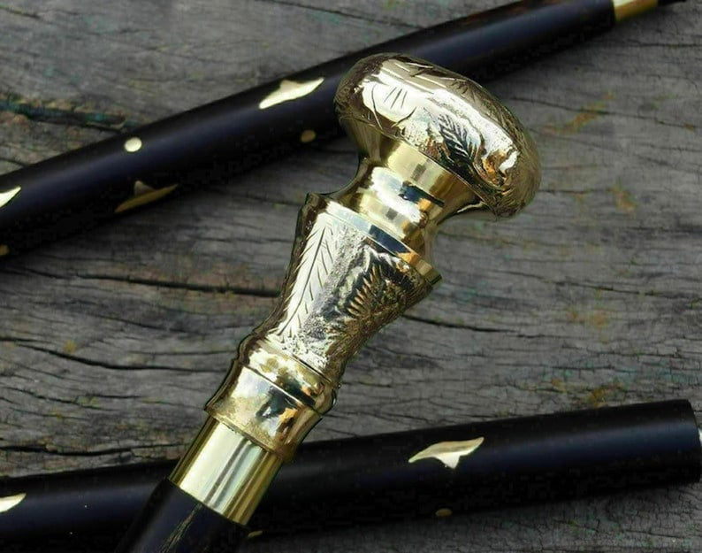 May include: A close-up of a black walking cane with a gold-colored handle. The handle has an ornate design with floral patterns. The cane's shaft is black with gold accents. The cane is resting on a wooden surface.