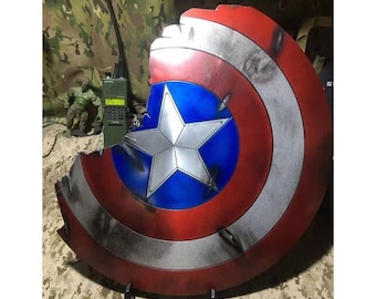 Peut inclure: Un bouclier de Captain America rouge, blanc et bleu avec une grande étoile argentée au centre. Le bouclier est fissuré et endommagé, avec un gros morceau manquant en haut. Le bouclier est présenté sur un support noir.
