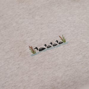 May include: A light gray sweatshirt featuring an embroidered design of a line of ducks swimming in a blue stream with green reeds. The ducks are black and white.
