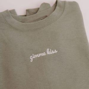 May include: Olive green sweatshirt with the embroidered text "gimme kiss" in white. The crew neck sweatshirt is folded, showing the neckline and part of the shoulder. The fabric appears soft and comfortable.