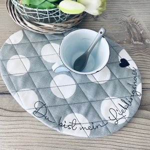 Può includere: Un sottobicchiere in tessuto grigio e bianco a pois con il testo "Du bist mein Lieblingshaus" ricamato in nero. Una tazza di smalto bianco con un cucchiaio è appoggiata sul sottobicchiere.