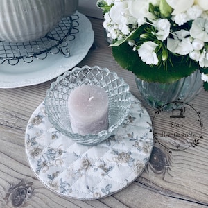 Könnte beinhalten: Eine graue Teekanne mit passendem Deckel steht auf einem weißen Teller mit einem Metall-Topfuntersetzer. Eine kleine Glasschale enthält eine Lavendelkerze, die auf einem Untersetzer mit floralem Muster ruht. Ein Strauß weißer Blumen befindet sich in einer Glasvase.