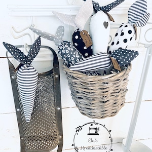 Könnte beinhalten: Eine Sammlung handgemachter Stoffkarotten in einem Weidenkorb. Die Karotten sind mit schwarzen und weißen Punkten, Streifen und Blumenmustern verziert. Die Karotten hängen an Haken an einer weißen Wand.