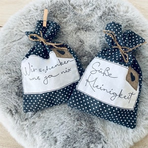 Könnte beinhalten: Zwei kleine Geschenktüten aus Stoff mit schwarzen und weißen Punkten und weißem Text. Der Text auf der linken Tüte lautet "Wir schenken uns ja nix". Der Text auf der rechten Tüte lautet "Süße Kleinigkeit".